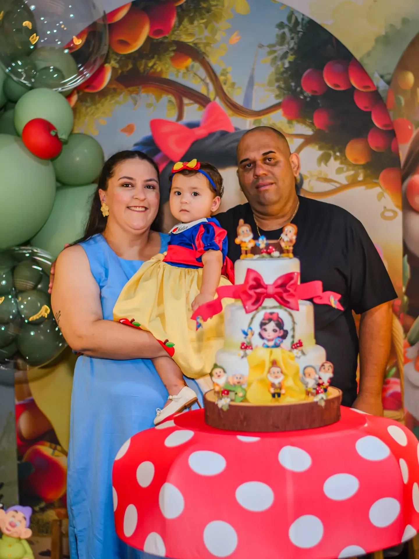 Criança entre duas pessoas diante de mesa de festa decorada.