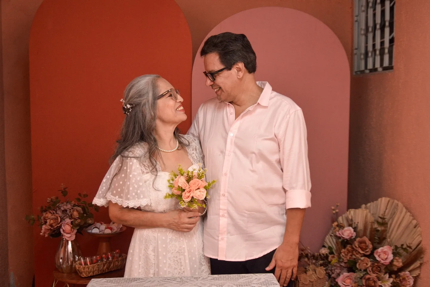 Casal com roupas de casamento diante de uma parede vermelha.