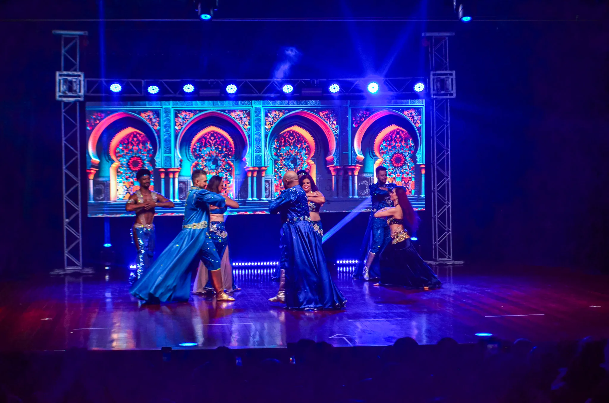 Grupo de dança com figurinos azuis e cenário de arcos coloridos.