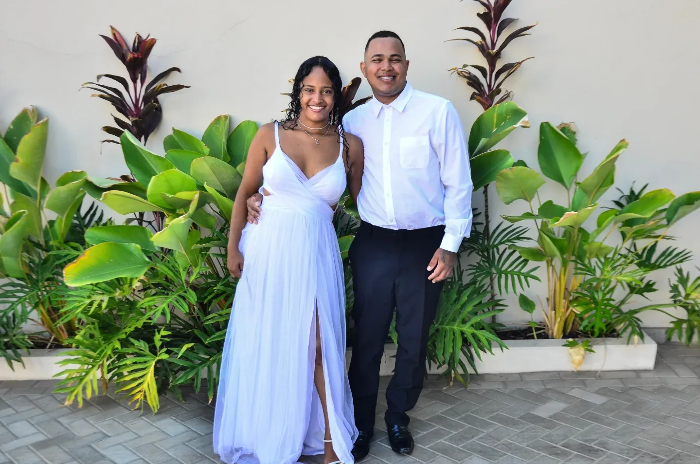 Casal com roupas de casamento diante de plantas e um muro.