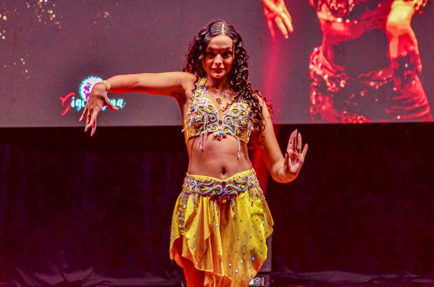 Dançarina com figurino dourado e os braços abertos no palco.