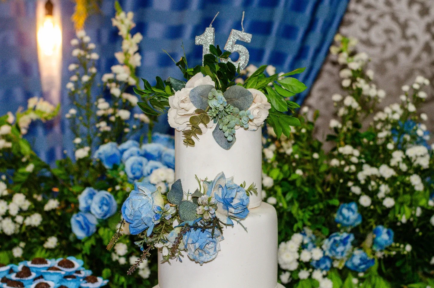 Bolo e flores diante de decoração com tecido azul.