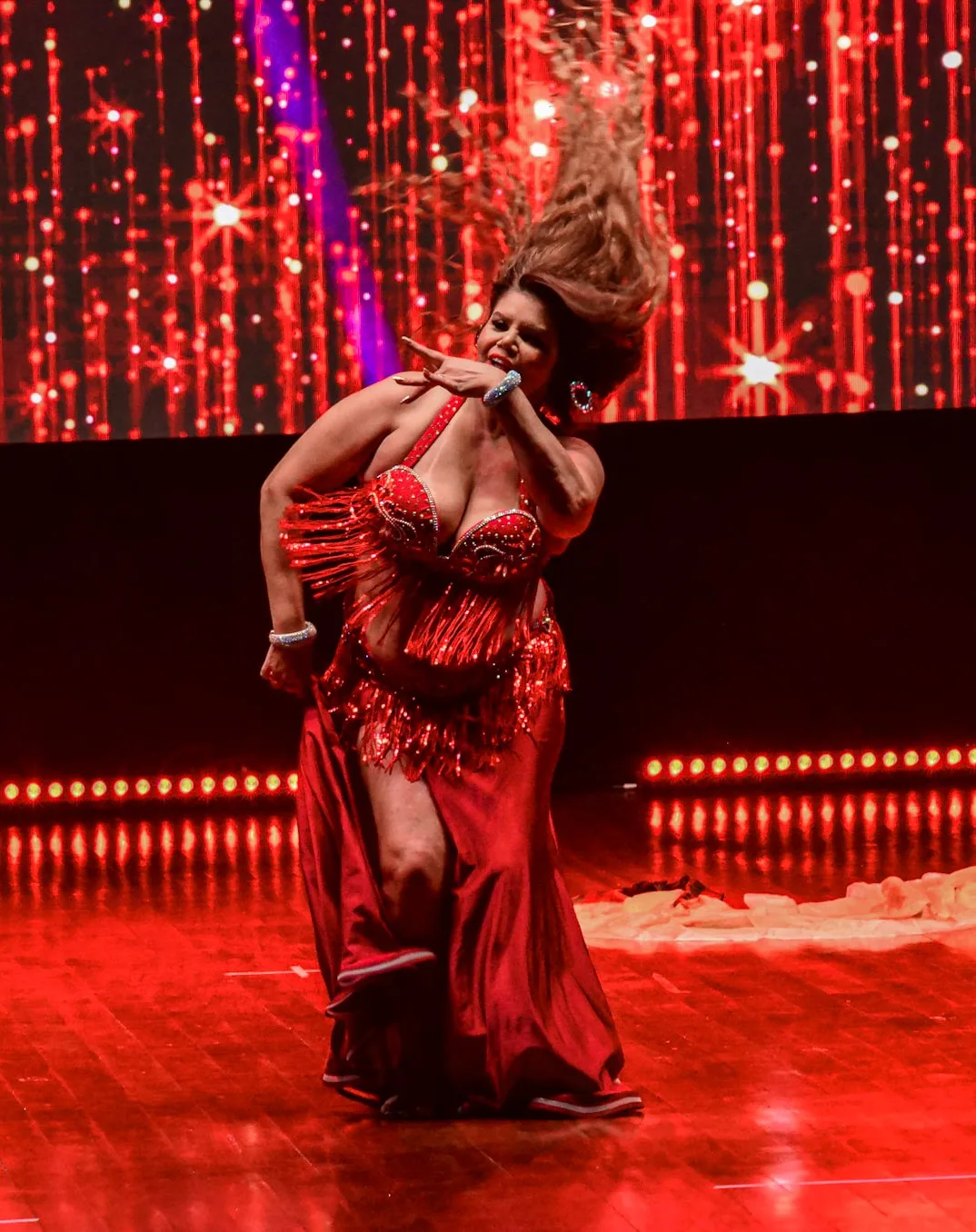 Dançarina em pose inclinada diante de cortina iluminada de vermelho.
